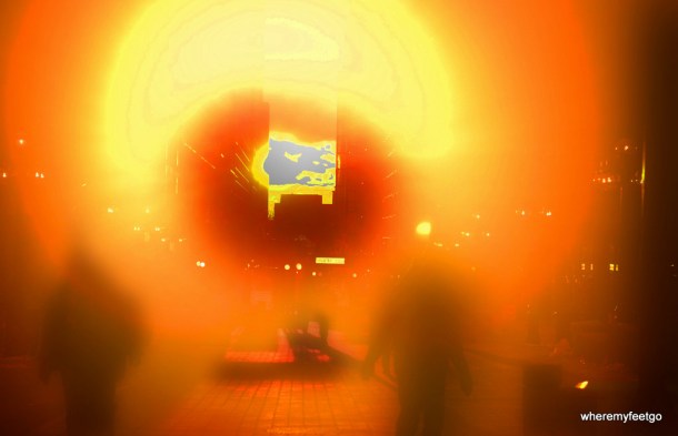 mostly yellow and orange light in a ring. there is a building in the distance in the centre of the light ring and two shadowy figures in the foreground.