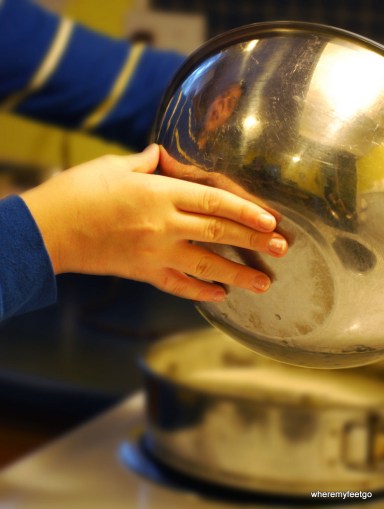 boy's face reflected in a metal mixing bowl. his hand is propping up the bowl like he is pouring something out of it.